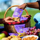 Person holding a package of 'Titi's Kitchen Chin Chin' with a picnic setup including fruits and cookies.